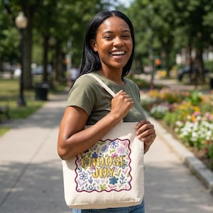 May include: A beige canvas tote bag featuring a scalloped-edge rectangular frame filled with a colorful floral pattern and the text CHOOSE JOY in yellow block letters. The bag has two fabric straps and is made of a light-colored woven material.