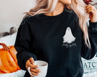 May include: Black sweatshirt with a white ghost graphic and the text "pretty spooky" on the front. The model is wearing blue jeans and holding a white mug.