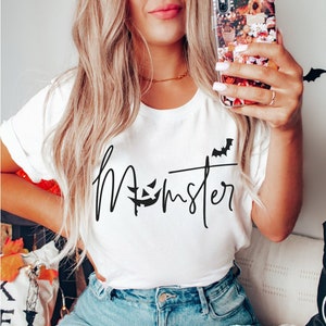 May include: A white t-shirt with the text "Momster" in black script with a jack-o-lantern face and a bat graphic.