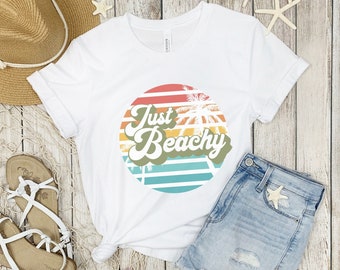 May include: White t-shirt with a retro sunset graphic and the text "Just Beachy". The graphic features palm trees and colorful stripes.