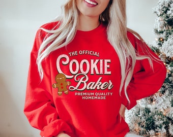 Könnte beinhalten: Roter Pullover mit einem Lebkuchenmann-Motiv und dem Text "The Official Cookie Baker Premium Quality Homemade".