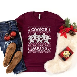 May include: A maroon t-shirt with a white Christmas sweater design featuring gingerbread men and the text "A Cookie Baking Crew".