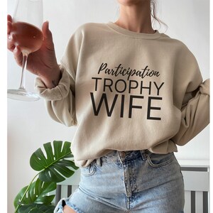 K&ouml;nnte beinhalten: Ein beiger Pullover mit dem Aufdruck "Participation Trophy Wife" in Schwarz.