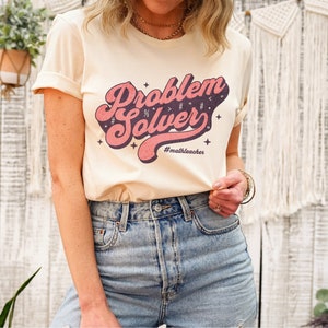 May include: A light beige t-shirt with a pink and purple retro-style graphic that says "Problem Solver" with the hashtag "#mathteacher" underneath.