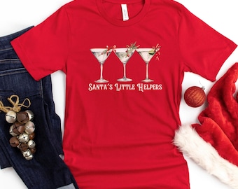 May include: A red t-shirt with the text "Santa's Little Helpers" and three martini glasses with festive garnishes. The shirt is paired with blue jeans, a Santa hat, and Christmas ornaments.