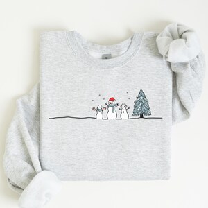 Op de afbeelding: Een lichtgrijs sweatshirt met een winterontwerp. De illustratie toont drie sneeuwpoppen, een kerstboom en vallende sneeuw. De sneeuwpoppen dragen een rode hoed, een blauwe sjaal en een blauwe hoed. Het sweatshirt is opgevouwen.