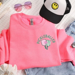 May include: Pink sweatshirt with a green and pink pickleball paddle graphic and the text "Pickleballer" on the front.