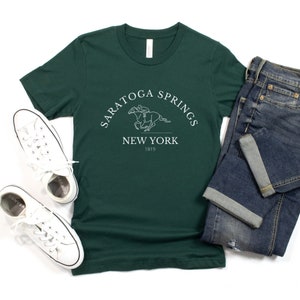 Puede incluir: Una camiseta verde oscuro con un dise&ntilde;o gr&aacute;fico blanco de un caballo y un jinete. El gr&aacute;fico incluye el texto "Saratoga Springs New York 1819".