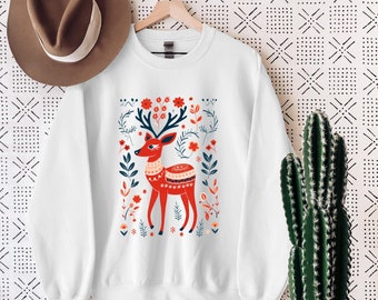 May include: A white crewneck sweatshirt featuring a central graphic of a red deer with dark blue antlers, surrounded by a pattern of red flowers and blue foliage. The deer has a decorative white and red patterned band across its body.