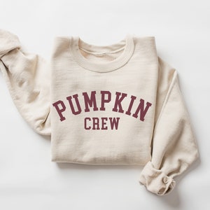 May include: A cream-colored sweatshirt with the words "PUMPKIN CREW" printed in maroon block letters. The sweatshirt is folded, with the sleeves rolled up. The crew neck sweatshirt is made of a soft material.