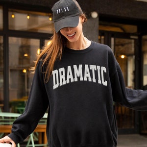 May include: Black sweatshirt with the word "DRAMATIC" printed in white on the front. The model is wearing a black baseball cap with the number "11:11" printed in white.