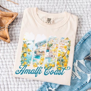 May include: A beige t-shirt with a watercolor image of the Amalfi Coast. The image is made up of four vertical panels, each showing a different view of the coastline. The panels are bordered by lemon trees with yellow lemons. The text 'Amalfi Coast' is printed in blue below the image.