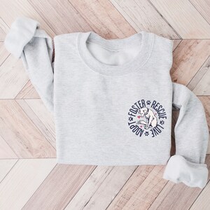 May include: A gray sweatshirt with a white dog illustration and the text "Adopt Foster Rescue Love" in a circle around the dog.