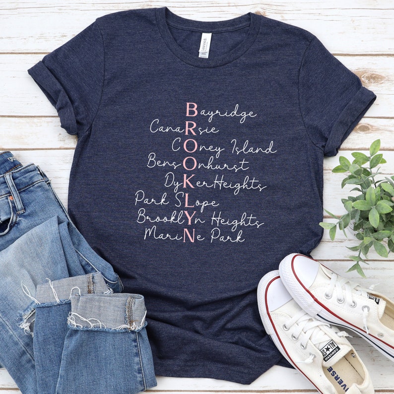 May include: A navy blue t-shirt with the word "BROOKLYN" spelled vertically in pink lettering. Each letter is a different neighborhood in Brooklyn, New York: Bayridge, Canarsie, Coney Island, Bensonhurst, Dyker Heights, Park Slope, Brooklyn Heights, and Marine Park.
