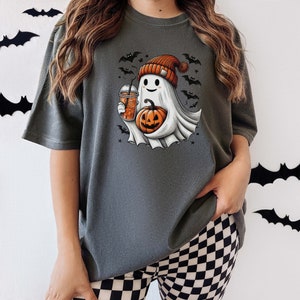 May include: A gray t-shirt with a cute ghost holding a pumpkin and a cup of iced coffee. The ghost is wearing a red and white hat with a pom-pom. The shirt has a Halloween theme with bats and stars printed on it.