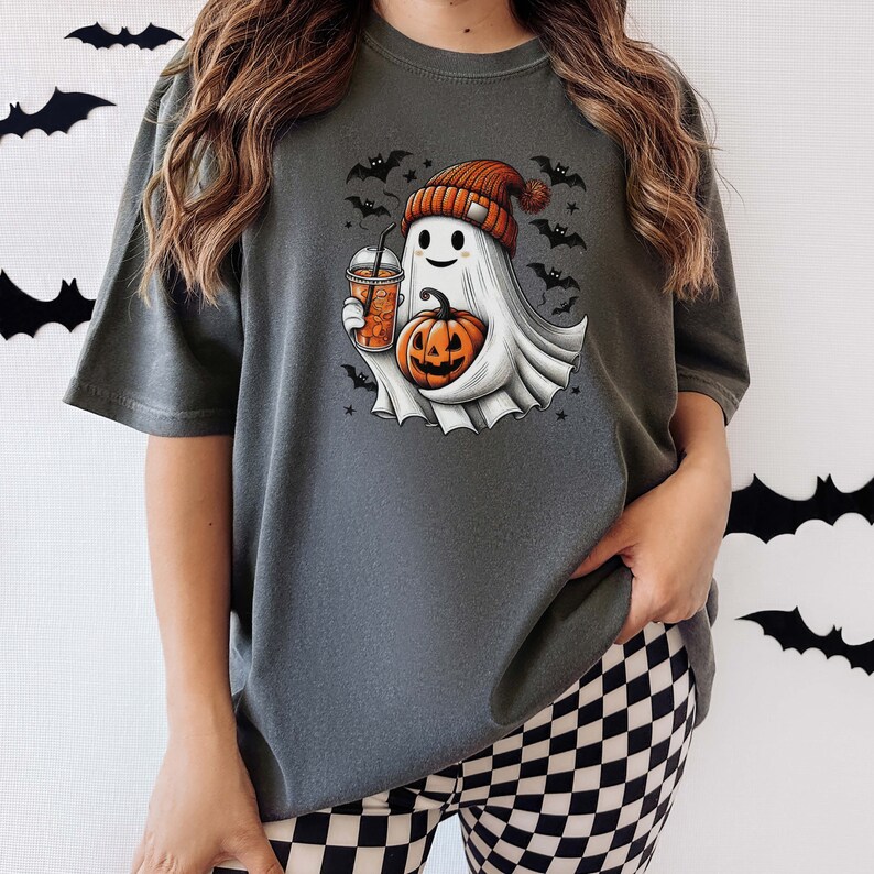 May include: A gray t-shirt with a cute ghost holding a pumpkin and a cup of iced coffee. The ghost is wearing a red and white hat with a pom-pom. The shirt has a Halloween theme with bats and stars printed on it.