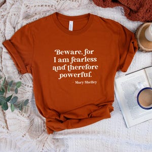 Puede incluir: Una camiseta de color &oacute;xido con una cita blanca que dice "Beware, for I am fearless and therefore powerful. - Mary Shelley". La camiseta est&aacute; sobre una manta blanca de ganchillo.