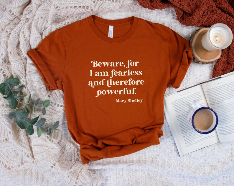 Puede incluir: Una camiseta de color &oacute;xido con una cita blanca que dice "Beware, for I am fearless and therefore powerful. - Mary Shelley". La camiseta est&aacute; sobre una manta blanca de ganchillo.