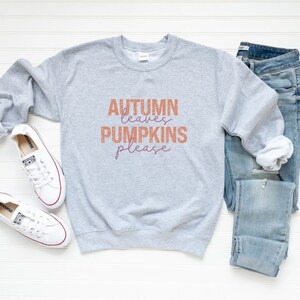 以下が含まれることがあります： オレンジと紫で「Autumn Leaves Pumpkins Please」の文字が入ったライトグレーのスウェットシャツ。秋をテーマにした、青いジーンズと赤いトリムの白いスニーカーも写っています。