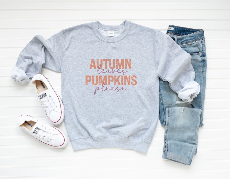 以下が含まれることがあります： オレンジと紫で「Autumn Leaves Pumpkins Please」の文字が入ったライトグレーのスウェットシャツ。秋をテーマにした、青いジーンズと赤いトリムの白いスニーカーも写っています。