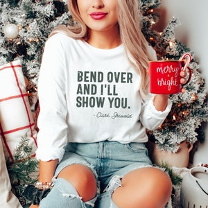 May include: White long-sleeved shirt with the text "Bend over and I'll show you. - Clark Griswold" printed on the front. A woman is wearing the shirt and holding a red mug with the text "merry + bright" printed on it.