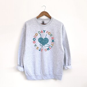 K&ouml;nnte beinhalten: Ein grauer Pullover mit einer bunten Grafik, auf der "Es ist ein guter Tag zum Stricken" steht, mit einem herzf&ouml;rmigen Wollkn&auml;uel und Stricknadeln.
