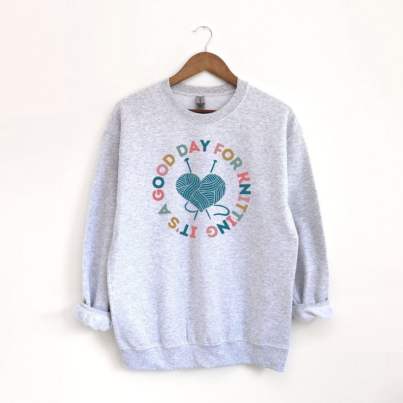 K&ouml;nnte beinhalten: Ein grauer Pullover mit einer bunten Grafik, auf der "Es ist ein guter Tag zum Stricken" steht, mit einem herzf&ouml;rmigen Wollkn&auml;uel und Stricknadeln.