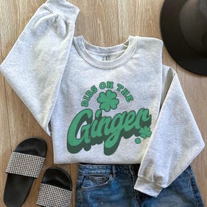 K&ouml;nnte beinhalten: Ein hellgraues Sweatshirt mit dem gr&uuml;nen Text "Dibs on the Ginger" und einem Kleeblatt. Das Sweatshirt wird mit Jeansshorts und schwarzen, strassbesetzten Pantoletten kombiniert. Im Hintergrund ist ein schwarzer Hut zu sehen.