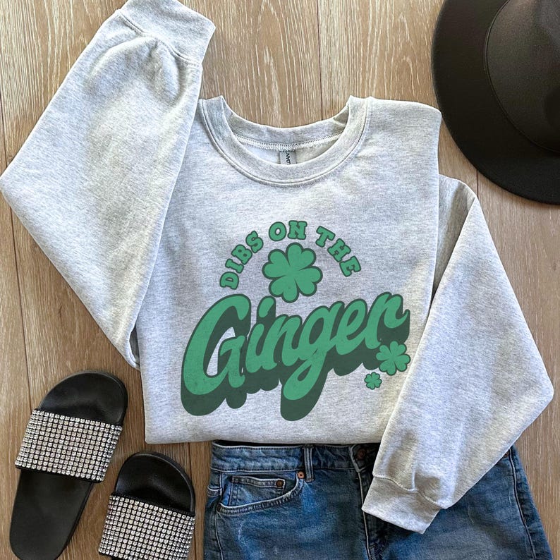 K&ouml;nnte beinhalten: Ein hellgraues Sweatshirt mit dem gr&uuml;nen Text "Dibs on the Ginger" und einem Kleeblatt. Das Sweatshirt wird mit Jeansshorts und schwarzen, strassbesetzten Pantoletten kombiniert. Im Hintergrund ist ein schwarzer Hut zu sehen.