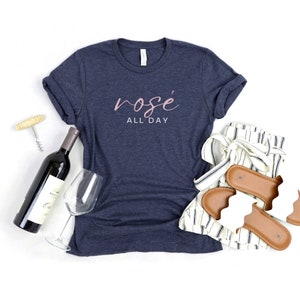 May include: A navy blue t-shirt with the text "ros&eacute; all day" in pink lettering. The shirt is laid flat on a white surface with a bottle of wine, a wine glass, a corkscrew, and a pair of white sandals with brown straps.