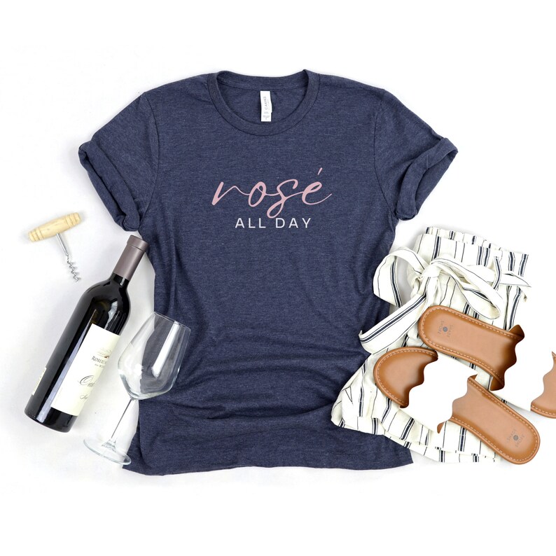 May include: A navy blue t-shirt with the text "ros&eacute; all day" in pink lettering. The shirt is laid flat on a white surface with a bottle of wine, a wine glass, a corkscrew, and a pair of white sandals with brown straps.