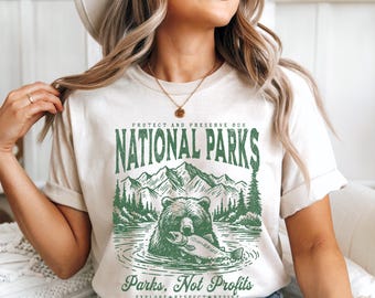 May include: A white t-shirt with a green graphic of a bear fishing in a lake with mountains and trees in the background. The text "Protect and Preserve Our National Parks" is above the graphic and "Parks, Not Profits" is below the graphic. The text "Explore. Respect. Resist." is below the bottom text.
