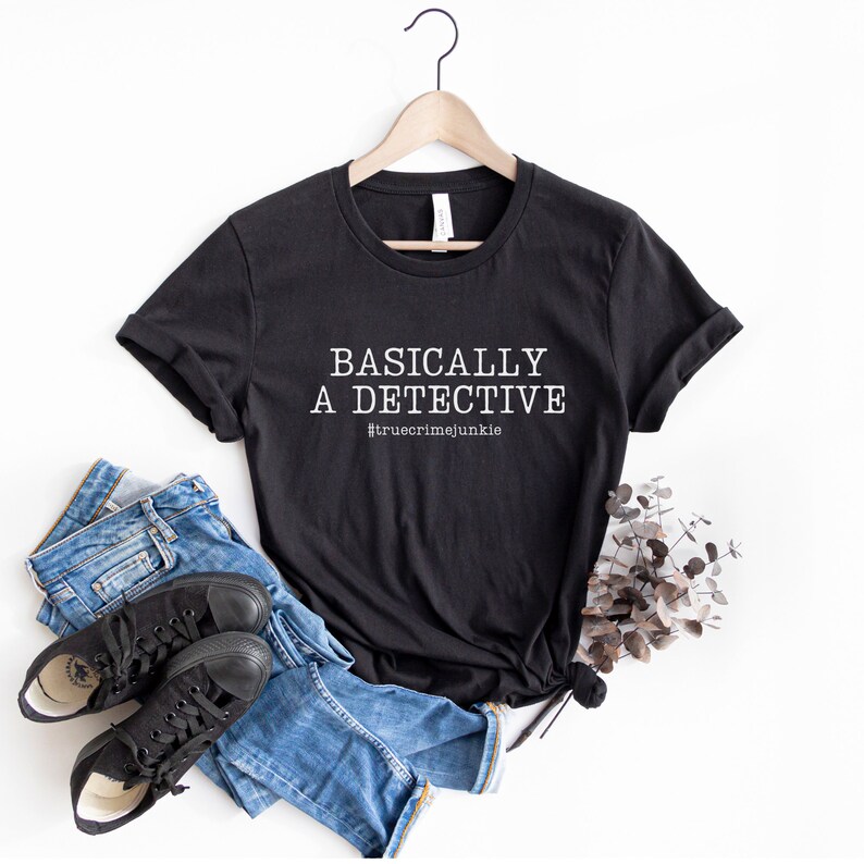May include: Black t-shirt with white text that reads "Basically a detective #truecrimejunkie". The shirt is on a hanger and there are jeans and black trainers in the image.