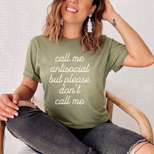 May include: A woman wearing a green t-shirt with the text "Call me antisocial but please don't call me" in white lettering. She is sitting in a wooden chair with her legs crossed.