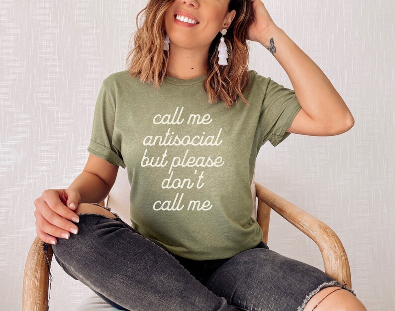 May include: A woman wearing a green t-shirt with the text "Call me antisocial but please don't call me" in white lettering. She is sitting in a wooden chair with her legs crossed.
