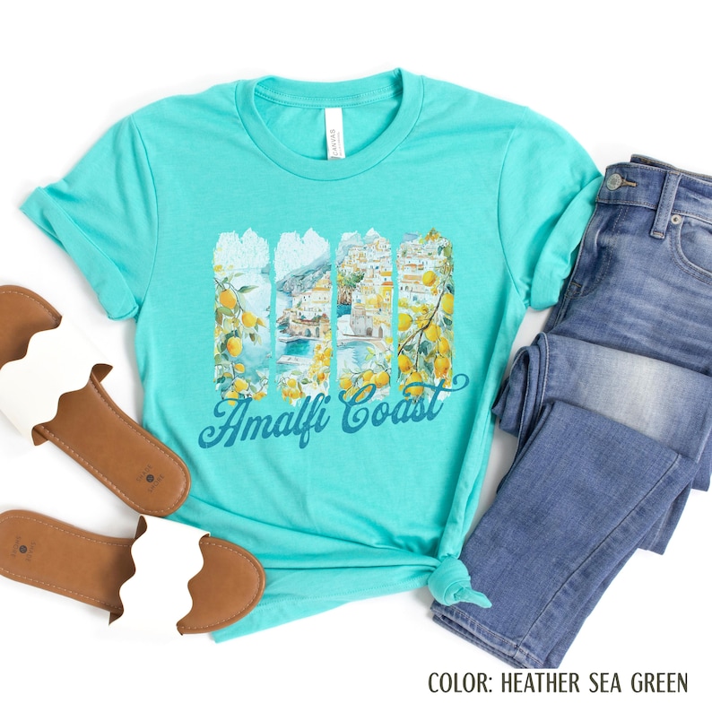 May include: A turquoise t-shirt with the words "Amalfi Coast" in a script font. The shirt features a graphic of the Amalfi Coast with lemons and buildings. The shirt is paired with blue jeans and sandals.