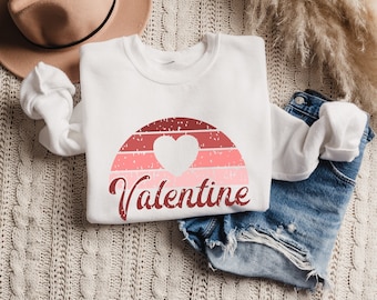 May include: White sweatshirt with a red and pink sunset graphic and the word "Valentine" in a red script font. A white heart is in the center of the sunset graphic.
