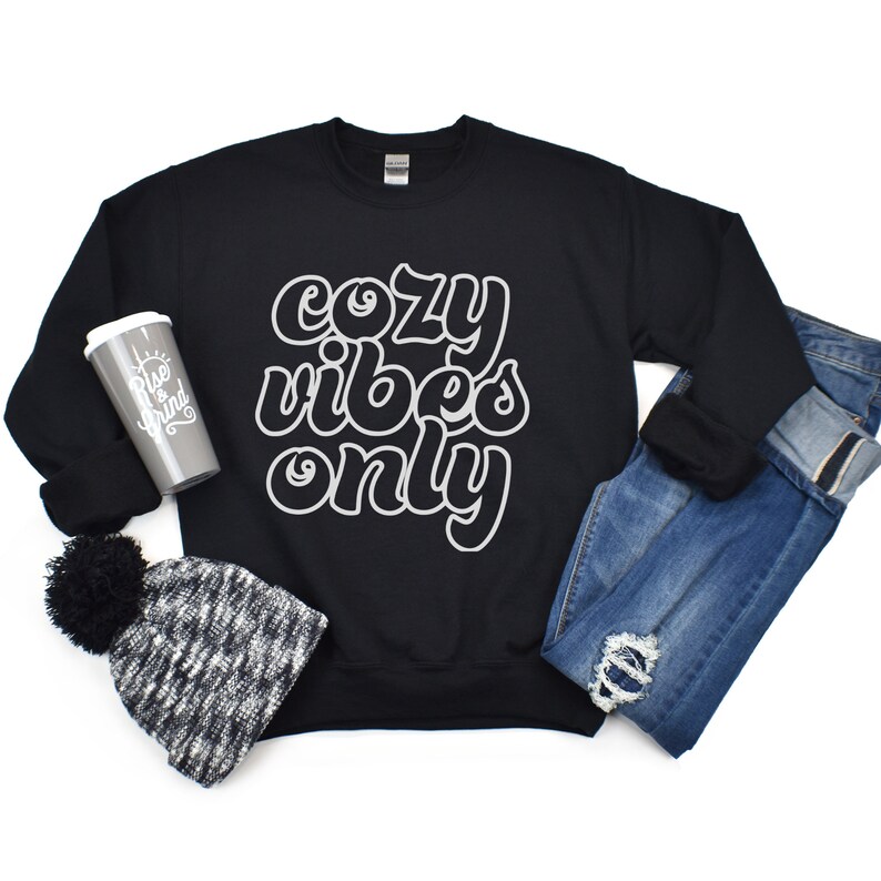 May include: Black sweatshirt with white text that reads "cozy vibes only". The sweatshirt is laid flat on a white background with a gray and white beanie and a gray travel mug.