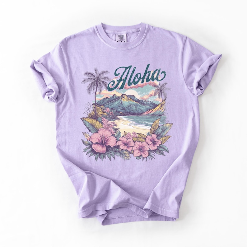 以下が含まれることがあります： ライラック色のTシャツ。トロピカルビーチのデザインで、ターコイズ色の筆記体で「Aloha」の文字、ヤシの木、山、ピンクのハイビスカスの花が描かれており、休暇をテーマにしています。
