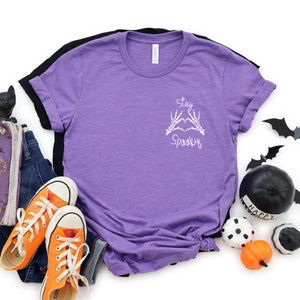May include: Purple short-sleeved t-shirt with a white graphic that says "Stay Spooky" with skeleton hands making a heart shape.