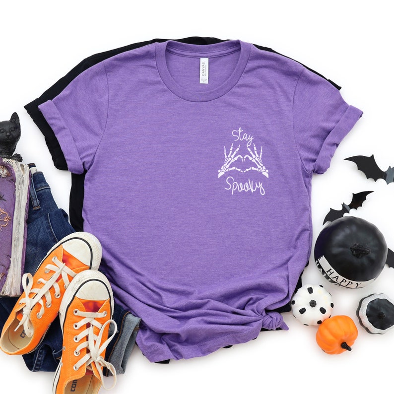 May include: Purple short-sleeved t-shirt with a white graphic that says "Stay Spooky" with skeleton hands making a heart shape.