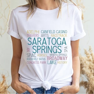 May include: White t-shirt with a teal and pink word cloud design that says "Saratoga Springs" with words like "Spa", "Horses", "Mineral Springs", "Broadway", "Congress Park", "Lake", and "History".