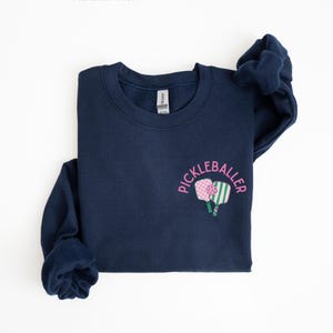 May include: A navy blue sweatshirt with the text "Pickleballer" in pink lettering and a graphic of two pickleball paddles.