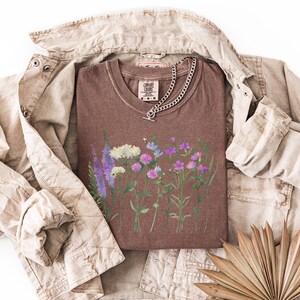 May include: A brown t-shirt featuring a watercolor floral design with purple, white, and pink flowers. A silver chain necklace with a toggle clasp rests on the shirt. The shirt is layered over a tan jacket.