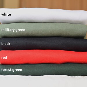 May include: A stack of five folded t-shirts in different colors: white, military green, black, red, and forest green.