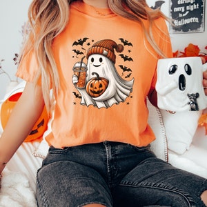 May include: Orange t-shirt with a cartoon ghost wearing a beanie hat and holding a pumpkin and a cup of iced coffee. The ghost is surrounded by bats and stars. The text "we're together, darling, every night is Halloween" is visible in the background.