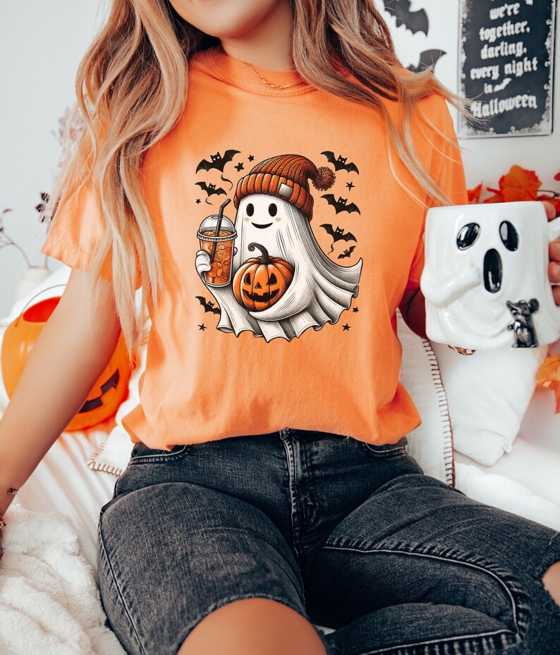 May include: Orange t-shirt with a cartoon ghost wearing a beanie hat and holding a pumpkin and a cup of iced coffee. The ghost is surrounded by bats and stars. The text "we're together, darling, every night is Halloween" is visible in the background.
