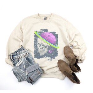 Może przedstawiać: Beżowa bluza z nadrukiem graficznym czaszki z planetą i pierścieniami wok&oacute;ł niej. Grafika jest w stylu vintage z ciemnoniebieskim tłem i gwiazdami.