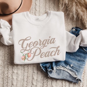 May include: White sweatshirt with the text "Georgia Peach" in brown script. A peach branch with two peaches is depicted next to the text.