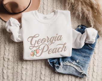 Könnte beinhalten: Weißer Pullover mit dem Text "Georgia Peach" in brauner Schrift. Neben dem Text ist ein Pfirsichzweig mit zwei Pfirsichen abgebildet.